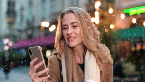 close-up view of caucasian woman using smartphone on the street while it‚äôs snowing in christmas