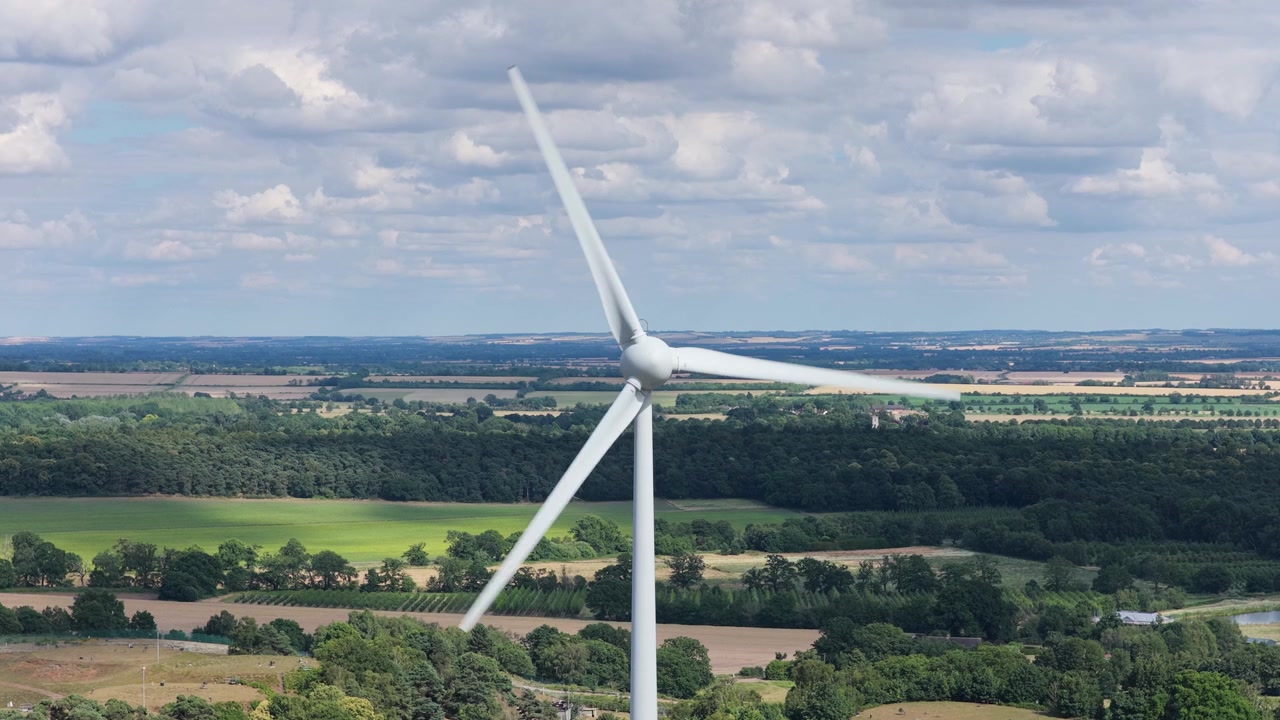 Pale bianche di turbina eolica ruotano lentamente contro un cielo azzurro brillante sopra campi verdi lussureggianti e natura rurale sotto la luce del sole durante un limpido pomeriggio estivo per la generazione di energia sostenibile