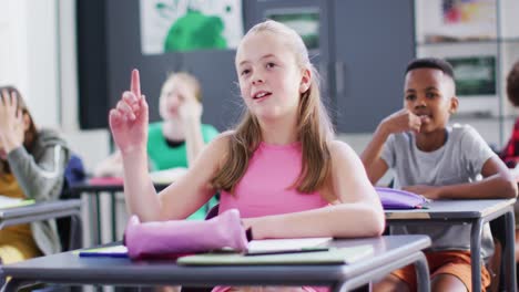 happy diverse schoolchildren at desks raising hands in school classroom