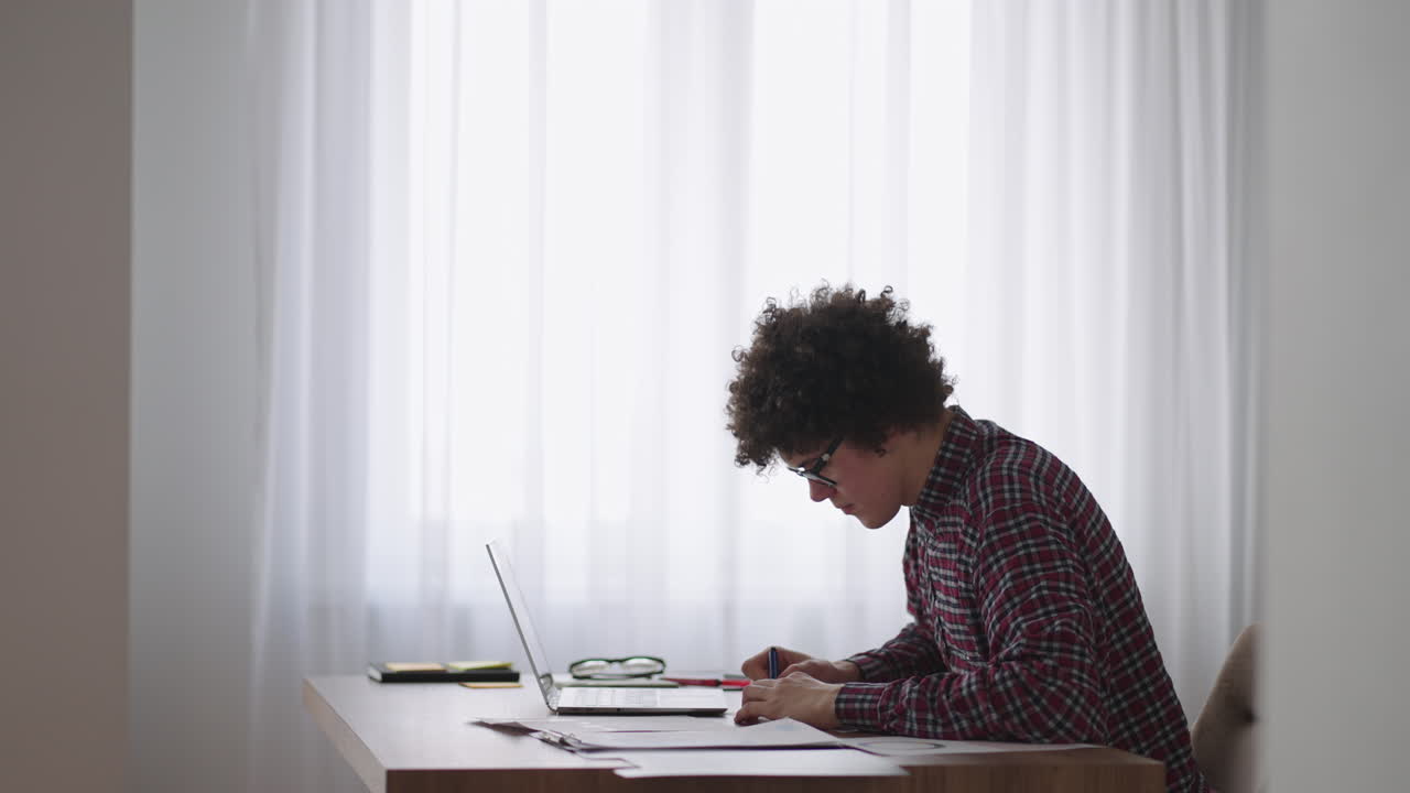 estudiante de cabello rizado atractivo joven con gafas está estudiando en casa usando una computadora portátil escribiendo en un cuaderno. estudiante universitario usando una computadora portátil viendo un seminario de aprendizaje en línea a distancia