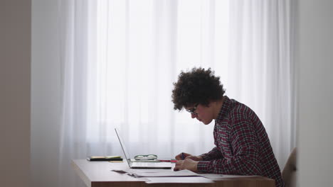 Estudiante-Masculino-De-Pelo-Rizado,-Un-Joven-Atractivo-Con-Gafas-Está-Estudiando-En-Casa-Usando-Una-Computadora-Portátil-Escribiendo-En-Un-Cuaderno.-Estudiante-Universitario-Usando-Una-Computadora-Portátil-Viendo-Un-Seminario-De-Aprendizaje-En-Línea-A-Distancia