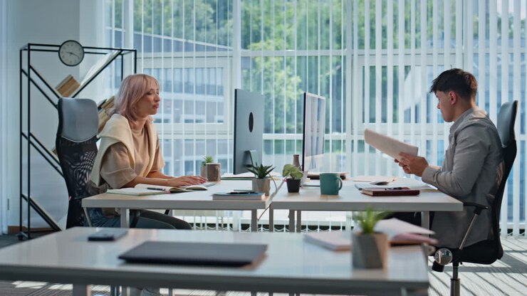 Donna che consegna dei documenti a un collega in un ufficio panoramico. Due colleghi che comunicano.