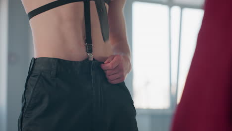 close up of bare body man holding black belt preparing to unfasten trousers while standing in front of person wearing red clothing during performance inside brightly lit studio space
