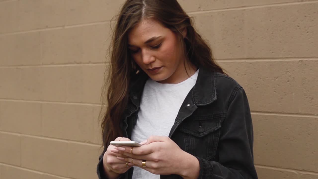 Steadicam shot of a beautiful,young, brunette college teenager texting on a cell phone wearing a white shirt with a black jacket against a wall