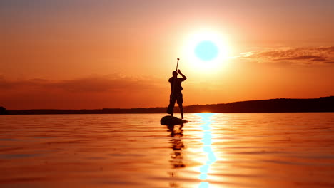 male silhouette rowing with an oar standing on a sup board. low angle view of a sportsman practicing and having fun at sunset.