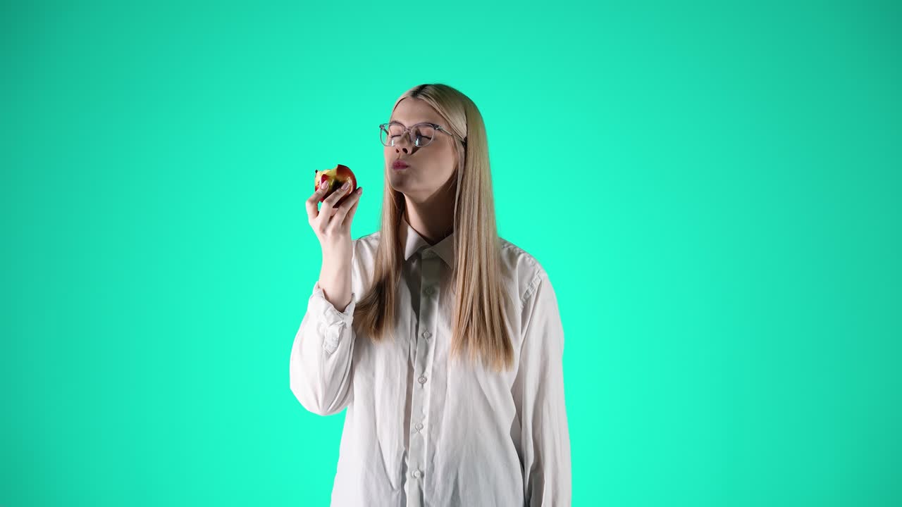Caucasian woman with glasses eating apple, taking a bite, turquoise background