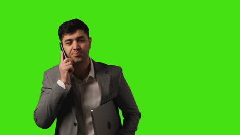 businessman with laptop talking on mobile phone walking into frame against green screen background