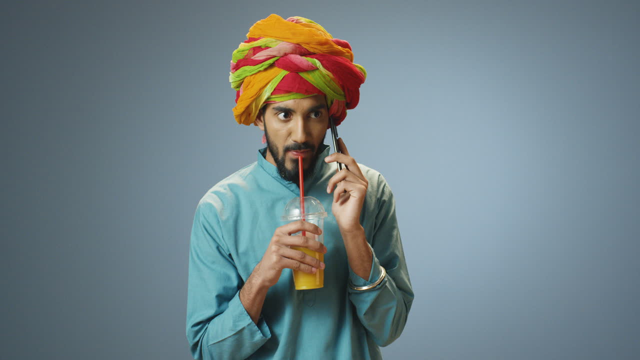 giovane uomo indiano allegro in abiti tradizionali e turbante che parla al telefono e beve succo d'arancia