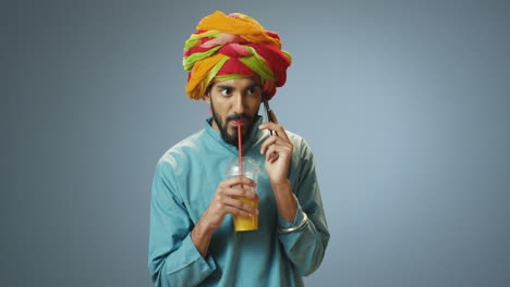 young cheerful indian man in traditional clothes and turban talking on the phone and drinking orange juice