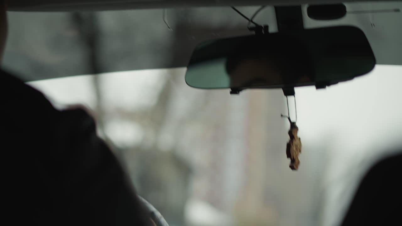 Rear view professional driver navigating inside car interior with center mirror swinging under overcast sky, steering wheel in hand showing skill control and concentration on road journey