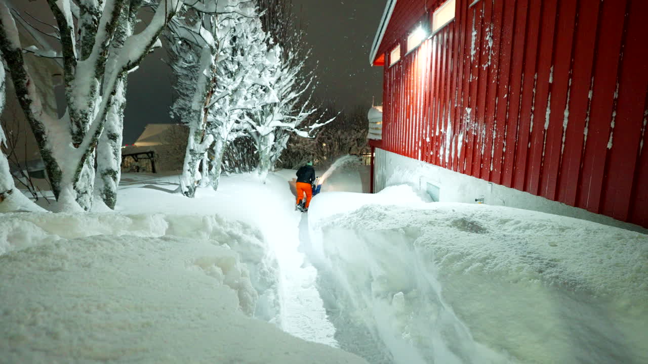 Rearview of man walking on snow covered Tromsø path clearing snow around building at night, arctic winter setting