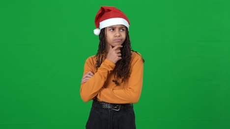 teen girl in new year hat thinking looking around choosing invisible christmas gifts on green screen. portrait of african american teenager posing at chromakey background. holidays season concept.
