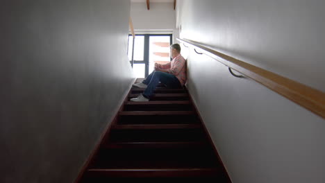 thoughtful senior biracial man sitting on stairs looking out of window, copy space, slow motion