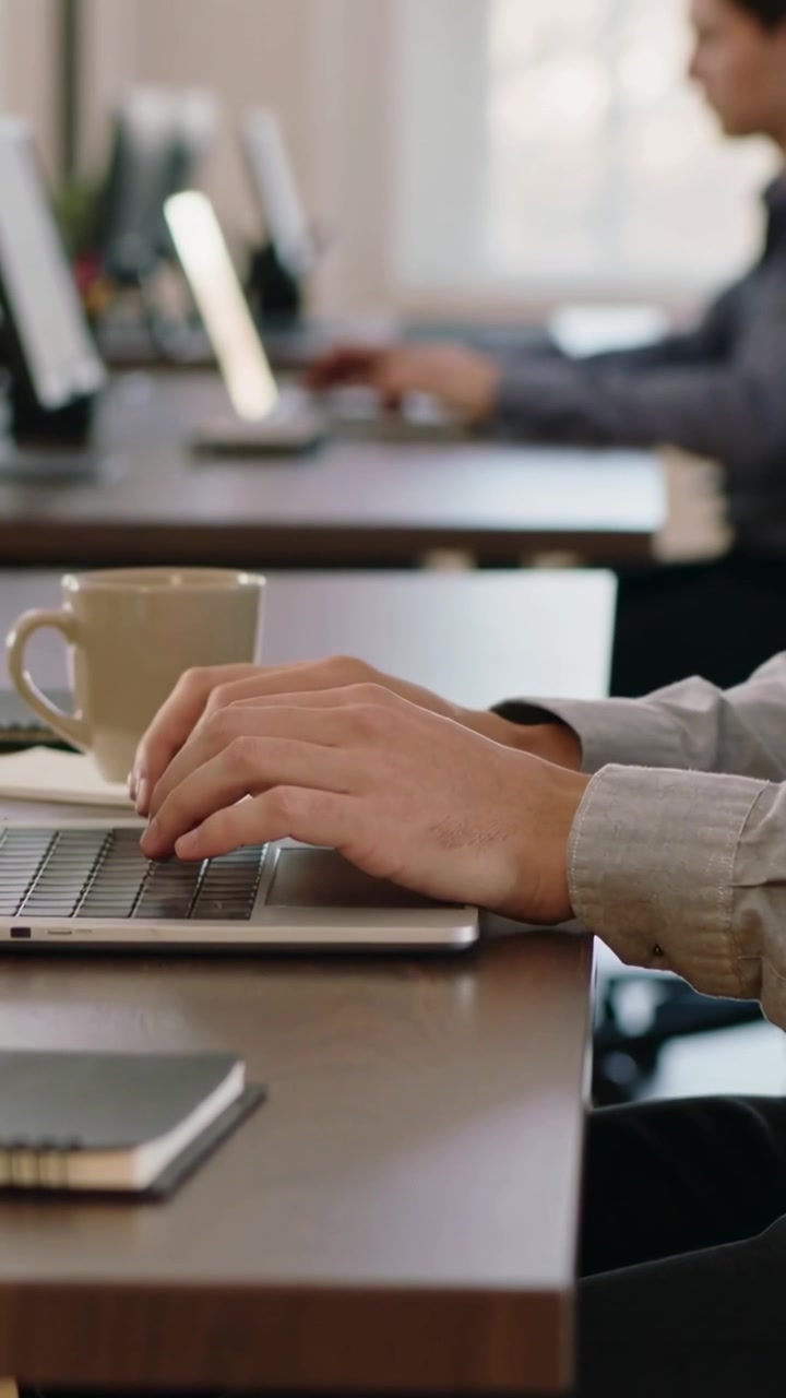 Video verticale: Inizio turno, mani con camicia chiara che digitano sul laptop alla scrivania, con tazza e telefono.