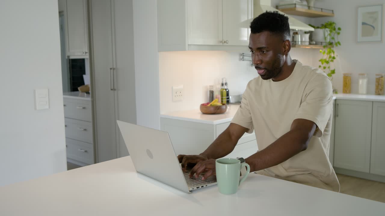 Adult man sitting at kitchen island typing on laptop, reaching for pale green mug drinking