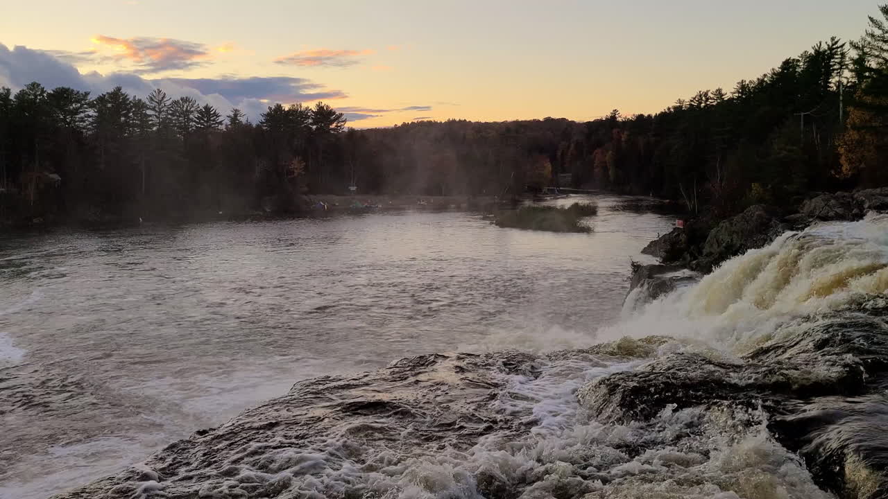A light mist forms in the morning air as a turbulent stream flows over rocks into a large river