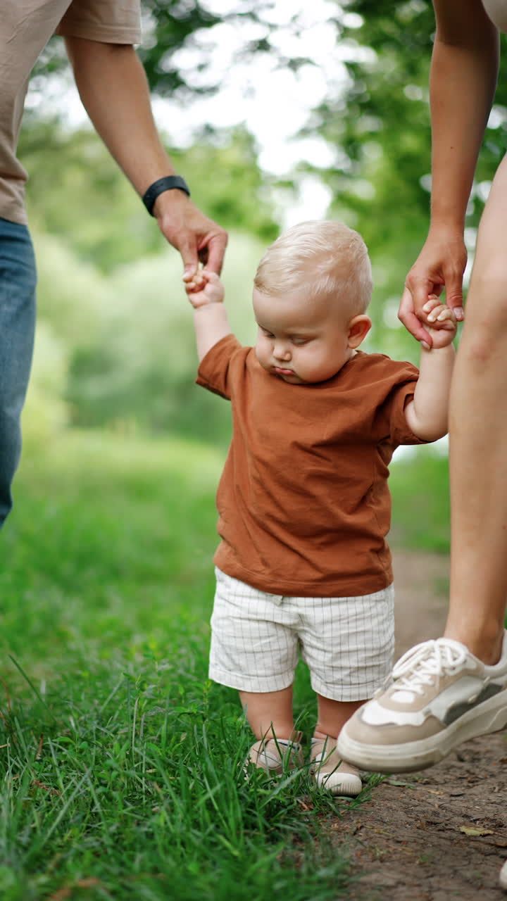 Unrecognized parents hold their baby by hands. Mom and dad dance but the kid watches them standing still. Blurred nature backdrop. Vertical video.