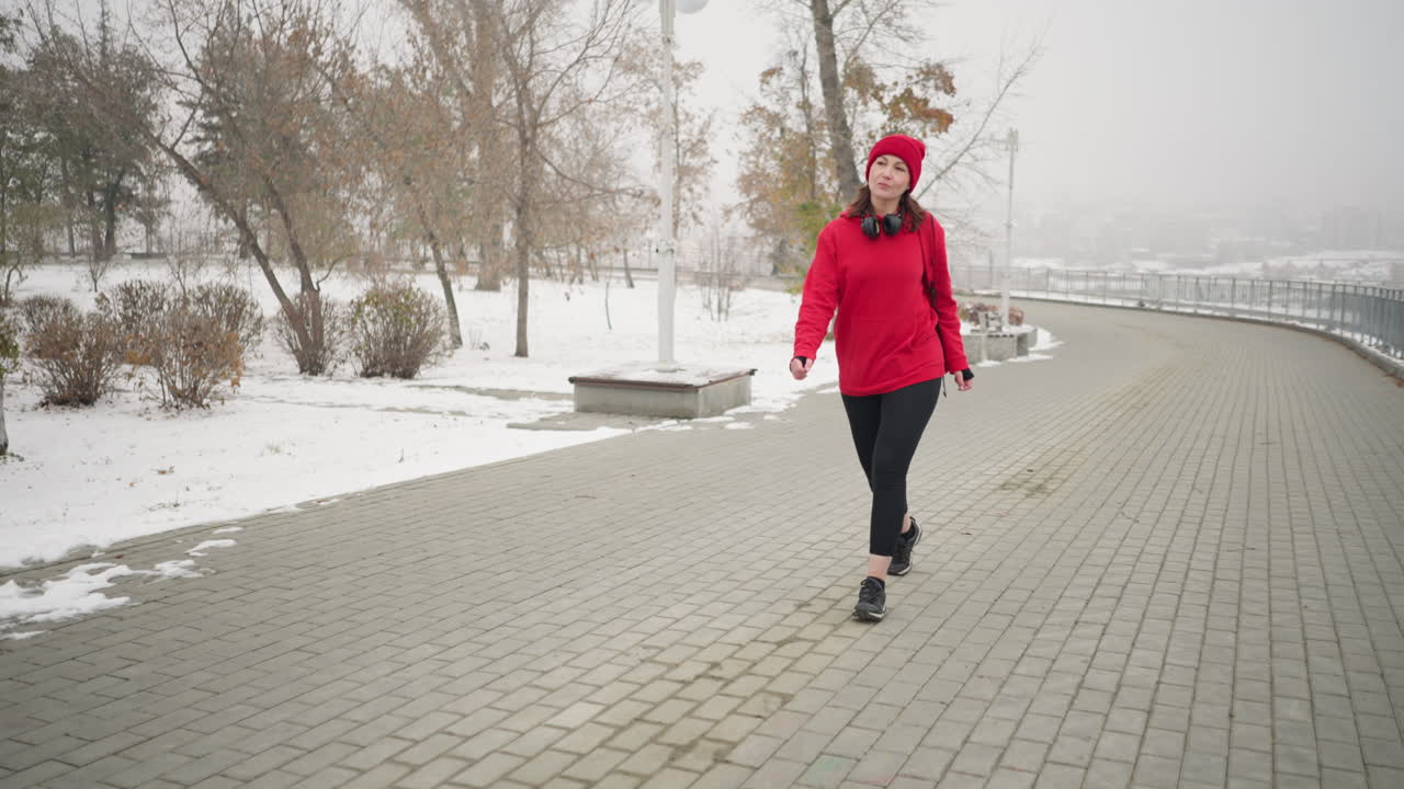 mujer de entrega con chaqueta roja y auriculares alrededor del cuello caminando por un camino entrelazado en un paisaje de parque nevado con árboles desnudos, arbustos, barandillas de hierro y postes de luz durante un día de invierno brumoso