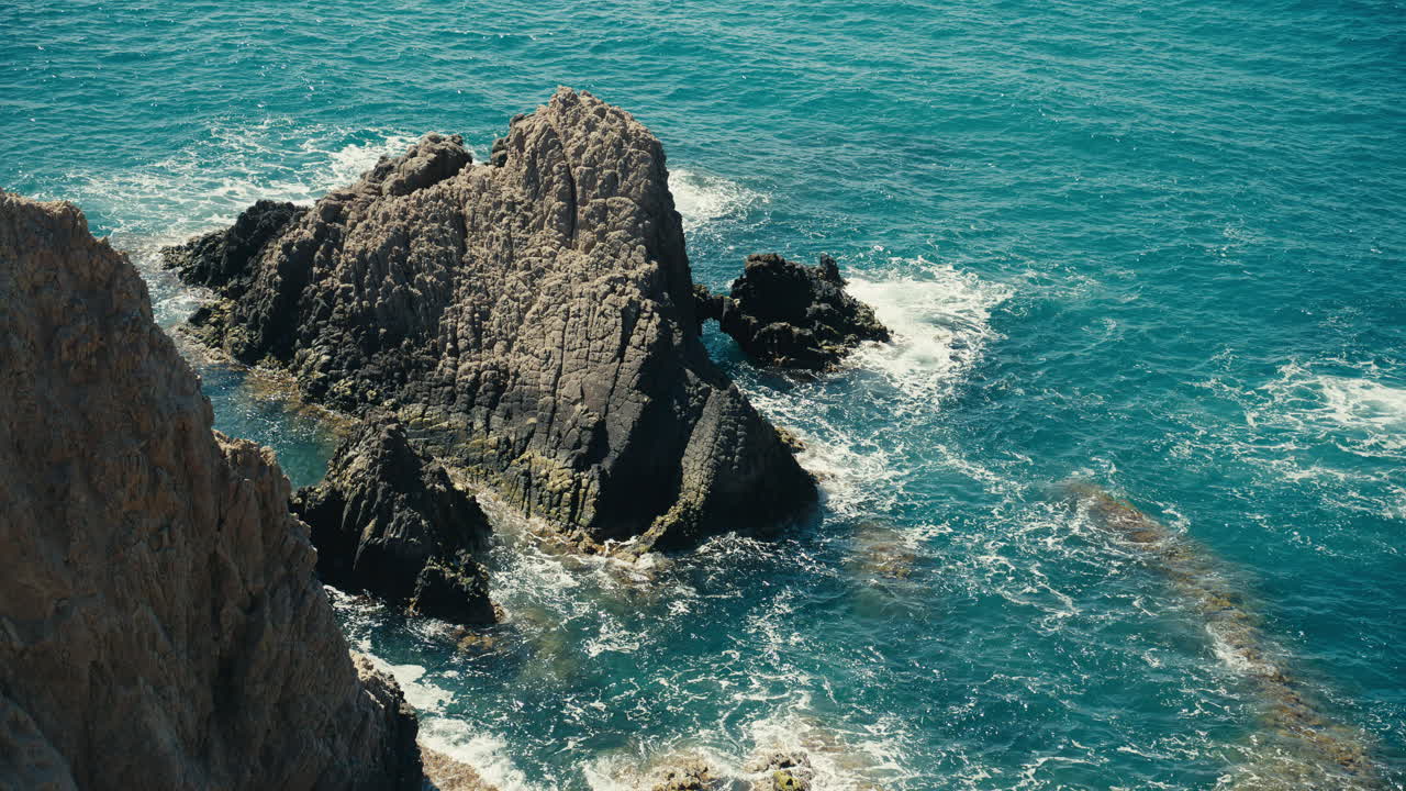 Dettagliata formazione rocciosa circondata da mare turchese lungo una costa frastagliata. Vista dall'alto che evidenzia le texture naturali, i modelli di erosione e le onde che si muovono attraverso un paesaggio marino mediterraneo.