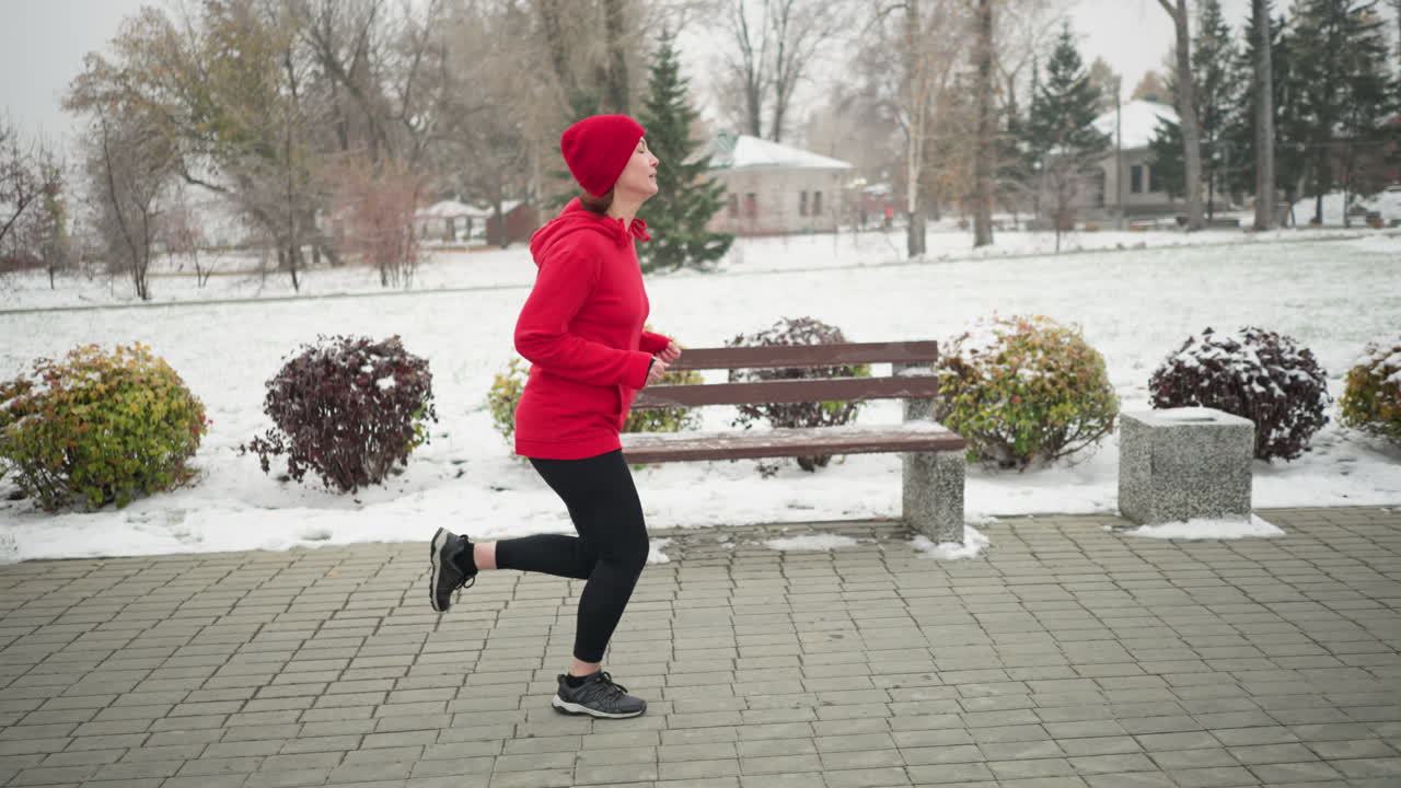 Side view of woman jogging along snowy park pathway in red hoodie and black leggings, surrounded by benches, colorful bushes, and frosty winter trees, with distant resident dusted with snow