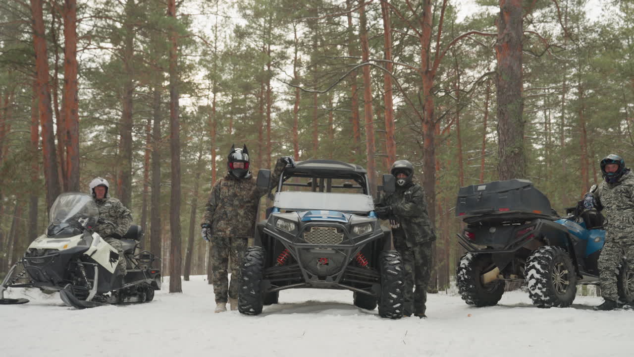 Schieramento di ATV e motoslitte nel bosco, equipaggio in tenuta mimetica pronto per una spedizione invernale estrema, caschi indossati, veicoli parcheggiati sulla neve, imponenti pini, fiocchi di neve che cadono, controlli pre-partenza e tensione.
