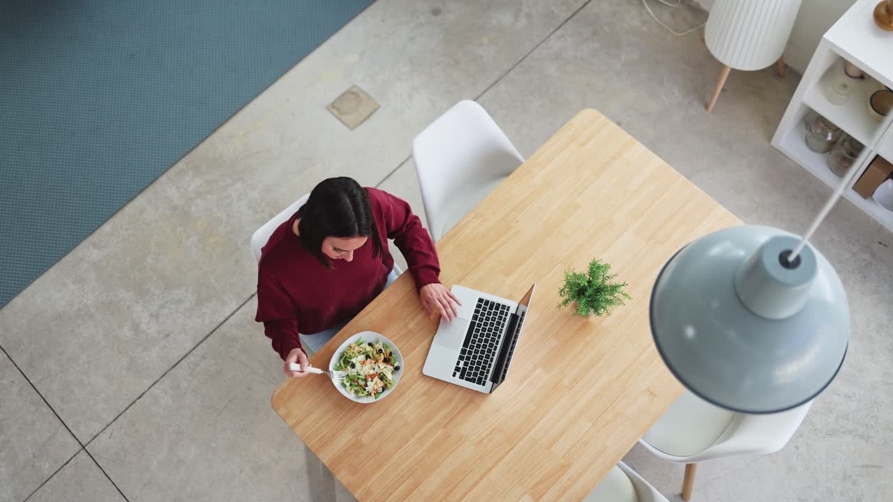 Vista dall'alto di una donna che mangia un'insalata e lavora al laptop a casa