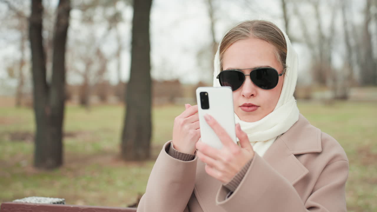 Dışarıda selfie çeken beyaz kadın, güneş gözlüğünü ayarlıyor ve çıplak ağaçların altında telefon kamerasıyla poz veriyor, şık başörtüsü ve kabanıyla neşeli bir sosyal ortamda içerik çeken bir influencer, bir blog yazarı.