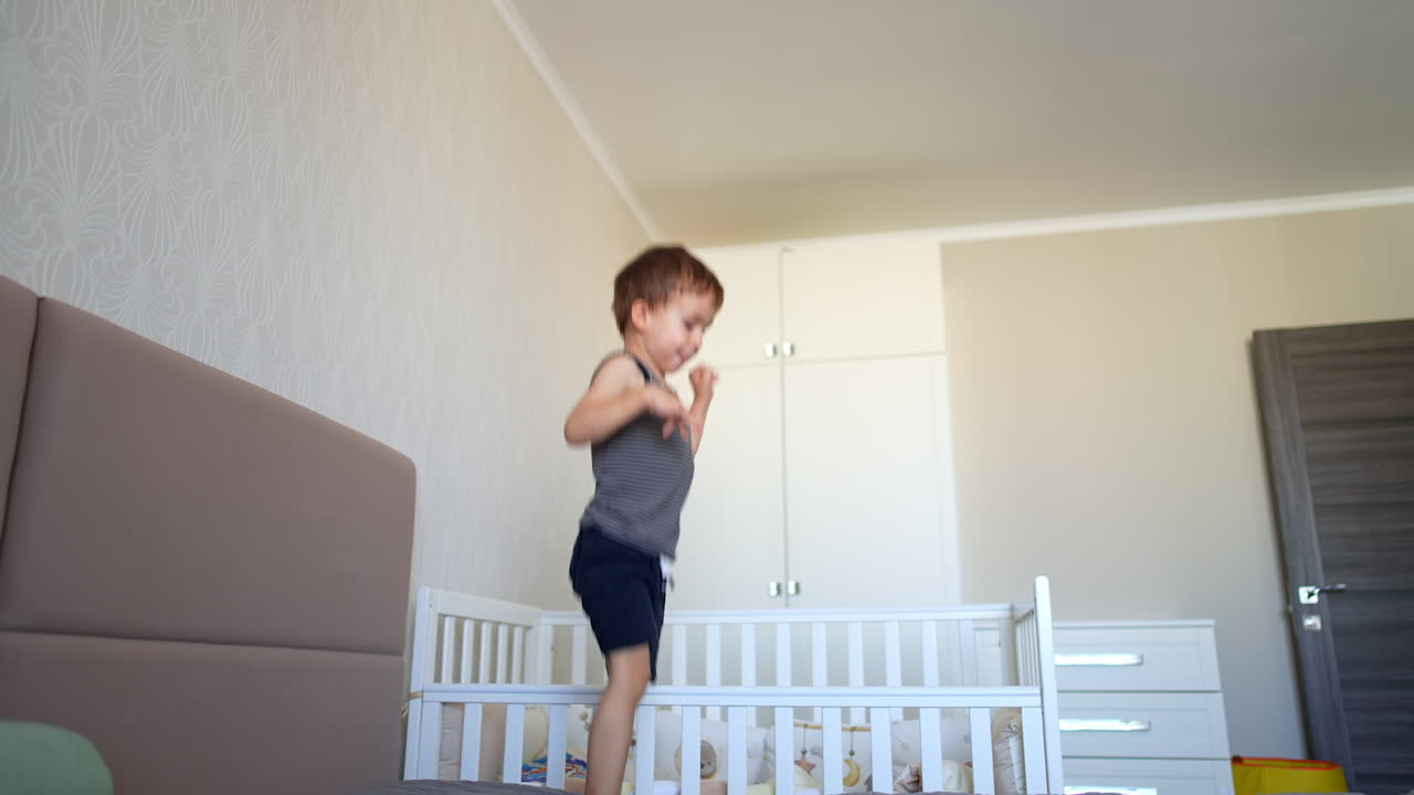 Cute toddler boy in sleeveless t-shirt and shorts bounces on the bed. Low angle view at the kid jumping in the bedroom.