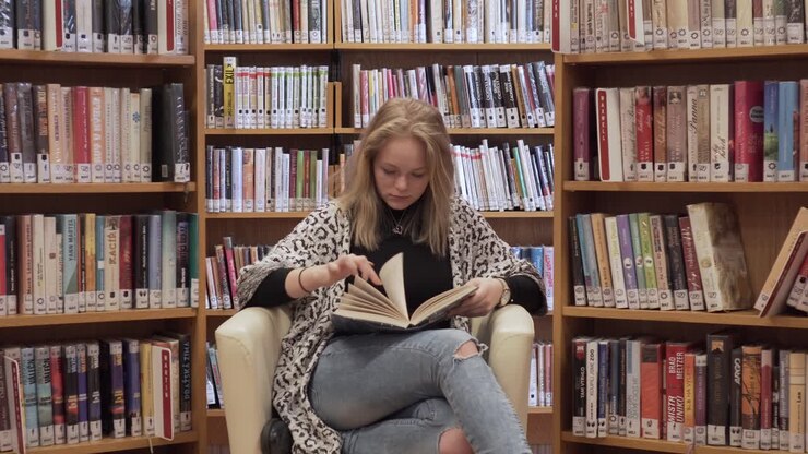 Studentessa seduta su una sedia che legge un libro con espressione seria, vista frontale.