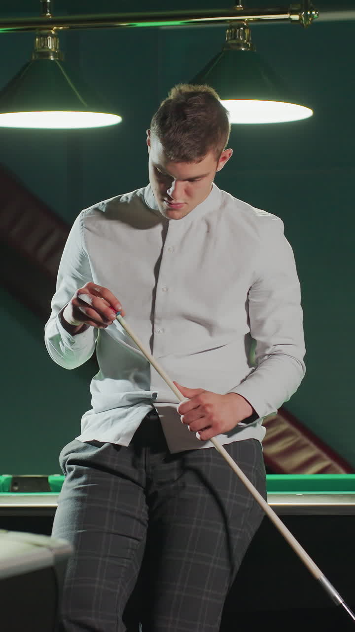 un joven jugador con camisa blanca y pantalones grises se sienta en la mesa de billar verde, sosteniendo el palo de la pilla con una expresión enfocada. otra mesa de billa es visible en el fondo bajo una iluminación tenue