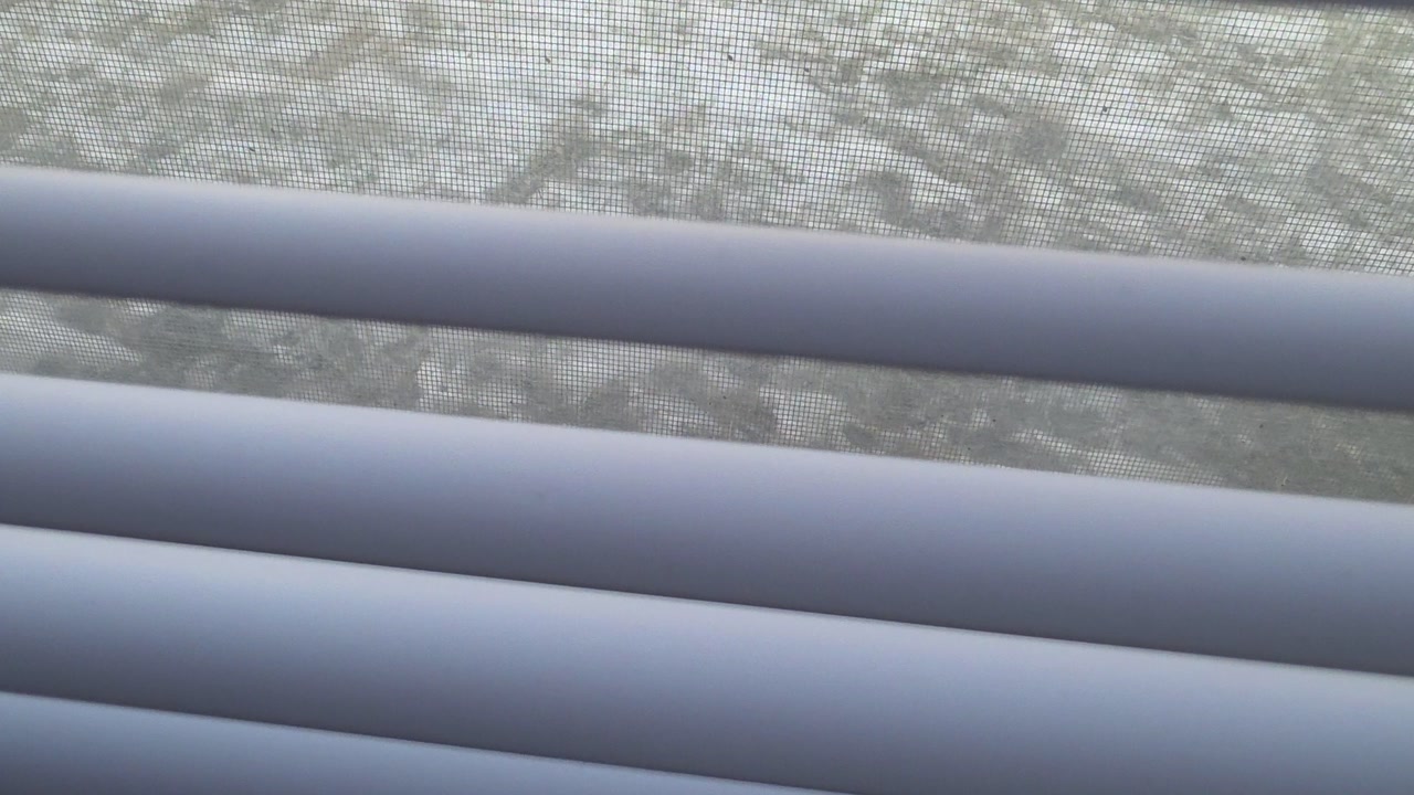 The camera glides upward, skimming the horizontal slats of clean white window blinds. Light filters through the narrow gaps, creating a rhythmic pattern of brightness and shadow as the view rises.