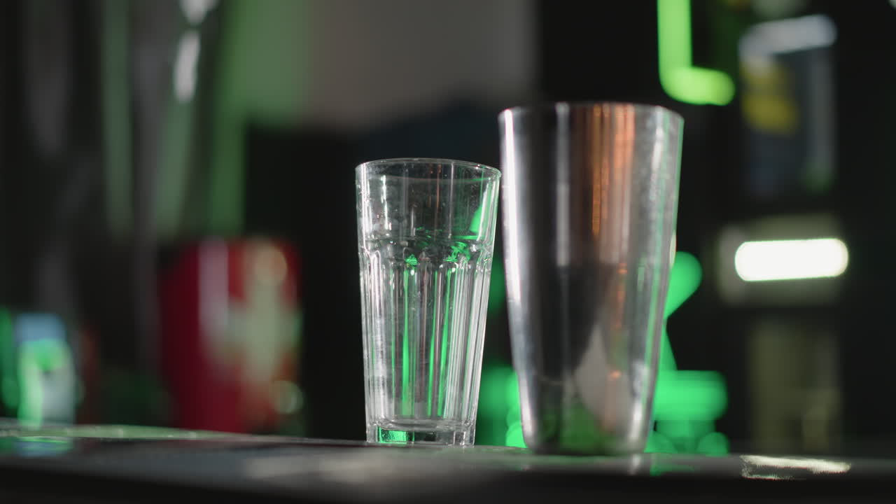 Close-up of empty glass and cocktail shaker on bar counter with vibrant green lighting in background, setting up for drink preparation in modern bar atmosphere