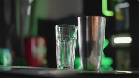 close-up of empty glass and cocktail shaker on bar counter with vibrant green lighting in background, setting up for drink preparation in modern bar atmosphere