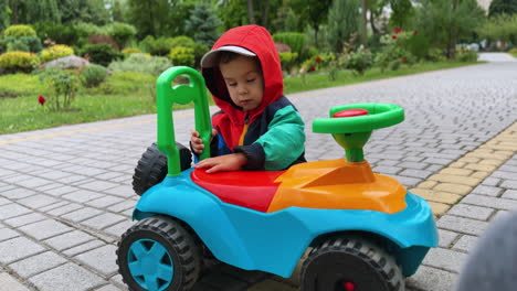 little baby boy sits near his colorful toy car. kid in a jacket with hood rises the seat of his car. park at backdrop.