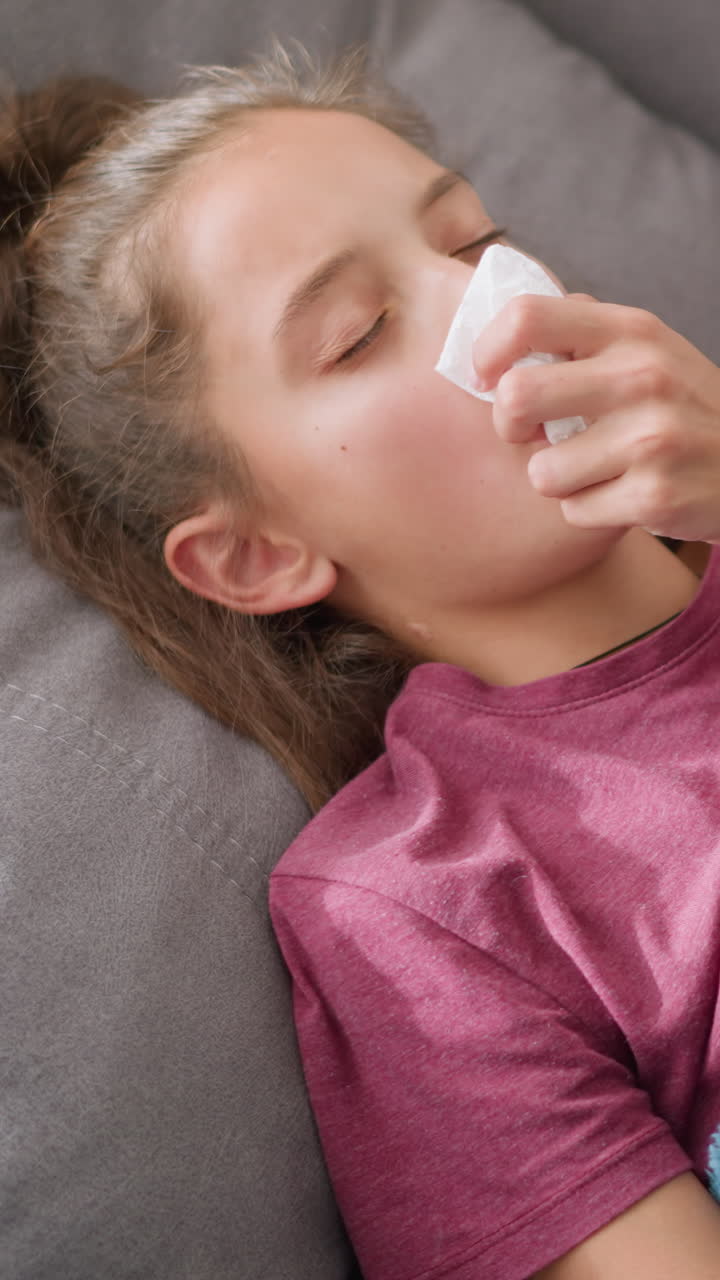 Aerial view of sick child lying on couch covered with soft blue blanket, holding tissue in left hand with red bangle, sneezing and resting, showing symptoms of cold or flu, feeling unwell