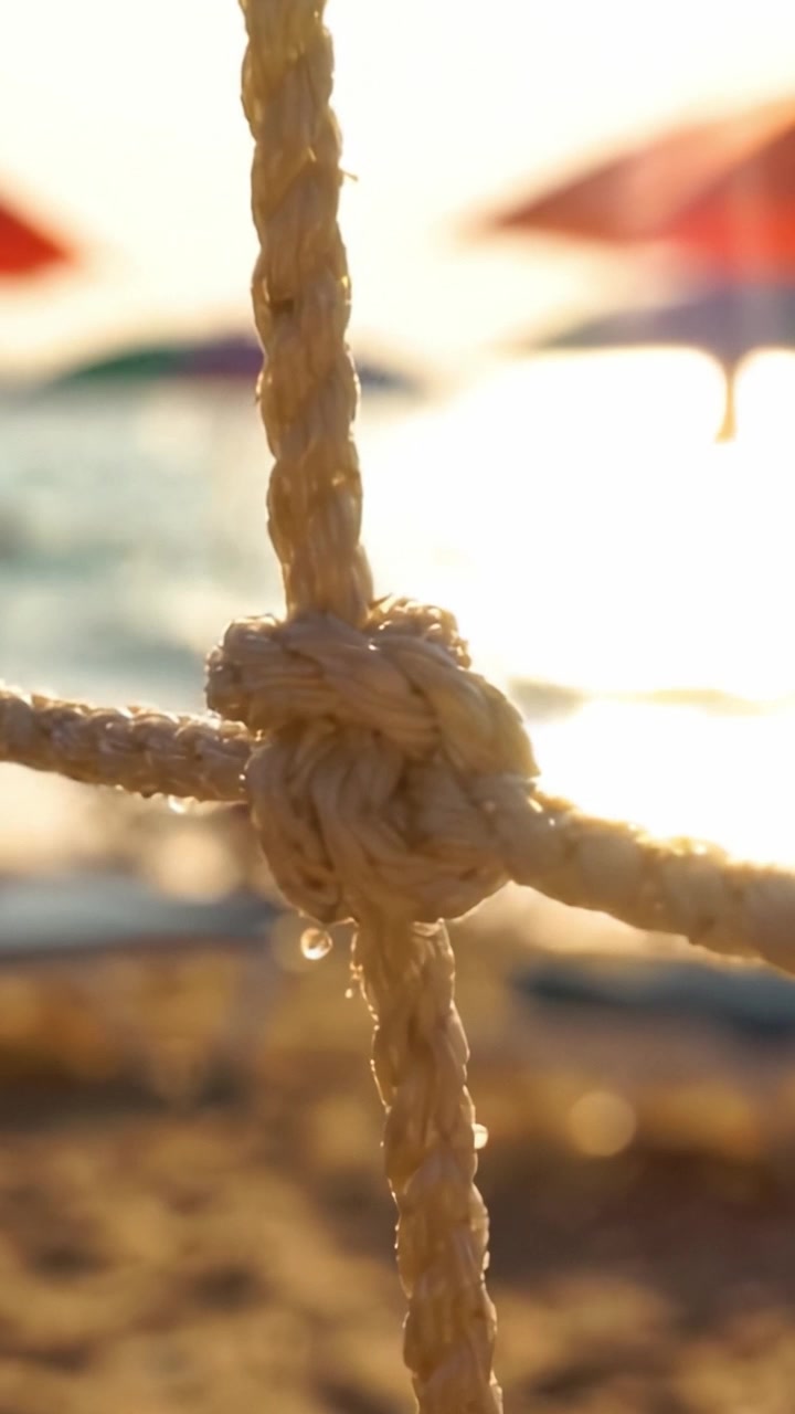 Primo piano di una corda annodata su uno sfondo di spiaggia vivido con luce solare e ombrelloni colorati, catturando l'essenza della tranquillità estiva.