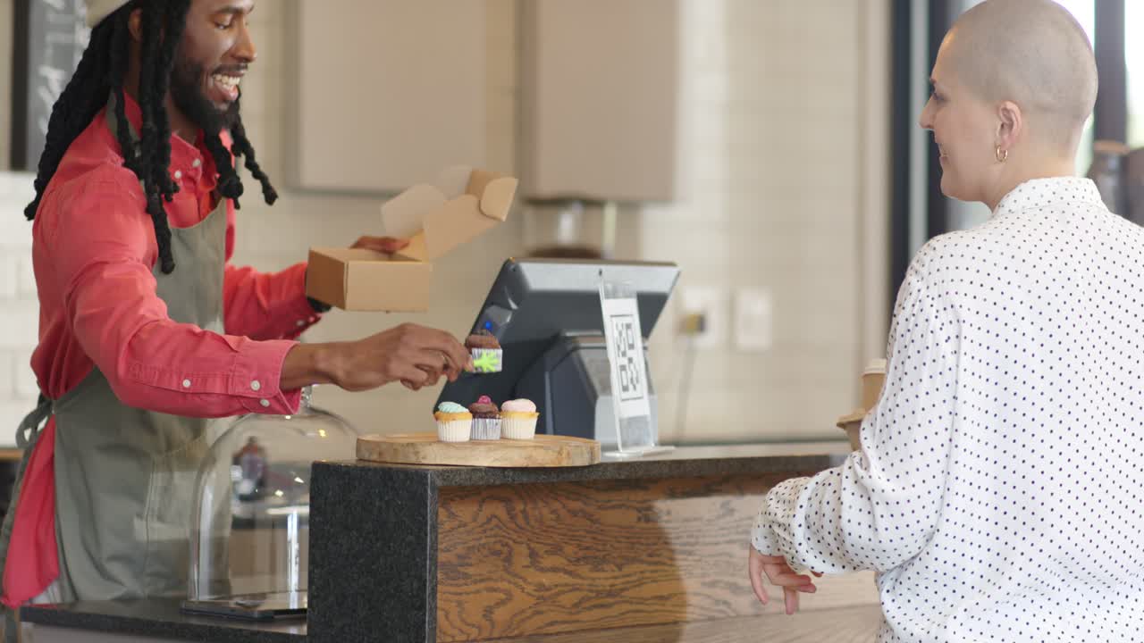 African American male barista boxing cupcakes in bakery as customer paying with QR code, copy space