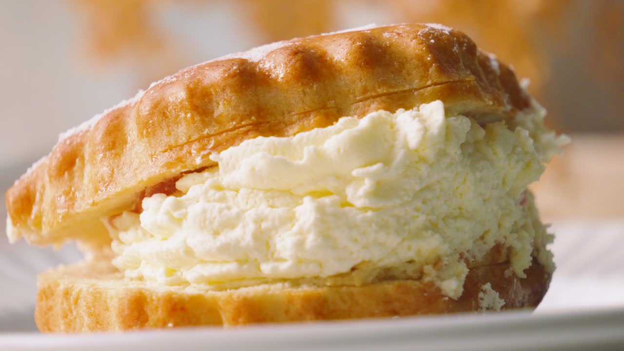 Slow Pan Across of Indulgent Cream and Jam Scone with Light Dusting of Icing Sugar on Top Resting on Plate in Natural Light. British Dessert from Devon and Cornwall. Classic Afternoon Tea.