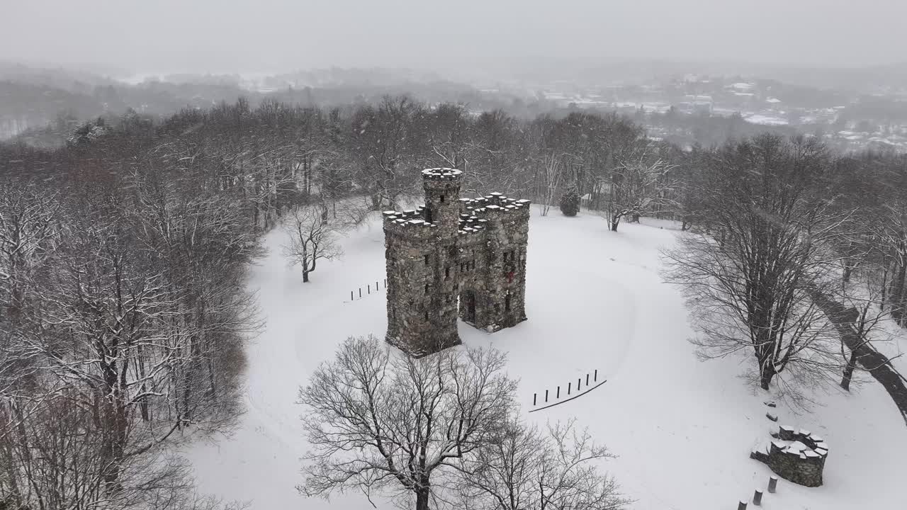Bancroft Tower sits amid snowy Worcester park. Stone fortress anchored in a white clearing. Overhead vantage reveals medieval-style architecture, aerial.