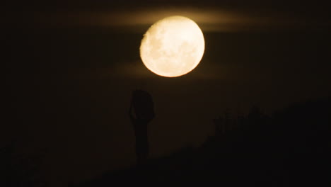 Silueta-De-Un-Surfista-Masculino-Sosteniendo-Una-Tabla-Larga-Contra-La-Luna-Llena-Con-Una-Sola-Aleta-Contra-La-Esquina-De-La-Luna-Levantando-La-Tabla-Sobre-La-Cabeza