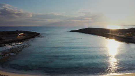 Aerial-shot-of-a-small-sandy-beach,-flying-over-a-single-swimmer-during-magic-hour