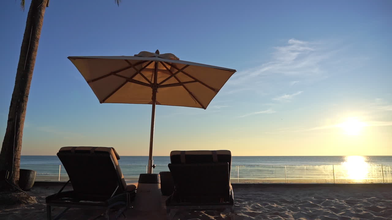 Two empty sun loungers and shade umbrella face a bright setting sun