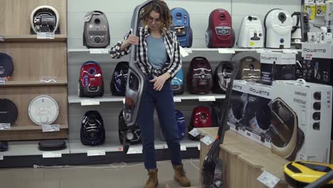 cheerful curly haired woman standing near new vacuum cleaners in home appliance store, choosing the right one. various vacuum cleaners in a row. slow motion