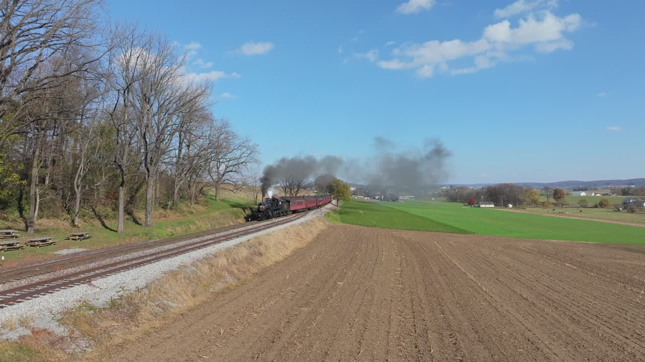 A classic retro steam train chugs along the tracks amid beautiful autumn scenery. The smoke billows from the engine as it moves past fields and trees on a clear, sunny day