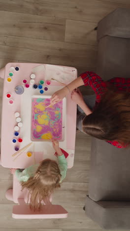 mother teaches little daughter to draw patterns on oily water surface at ebru art in living room upper view. creative lesson for children development