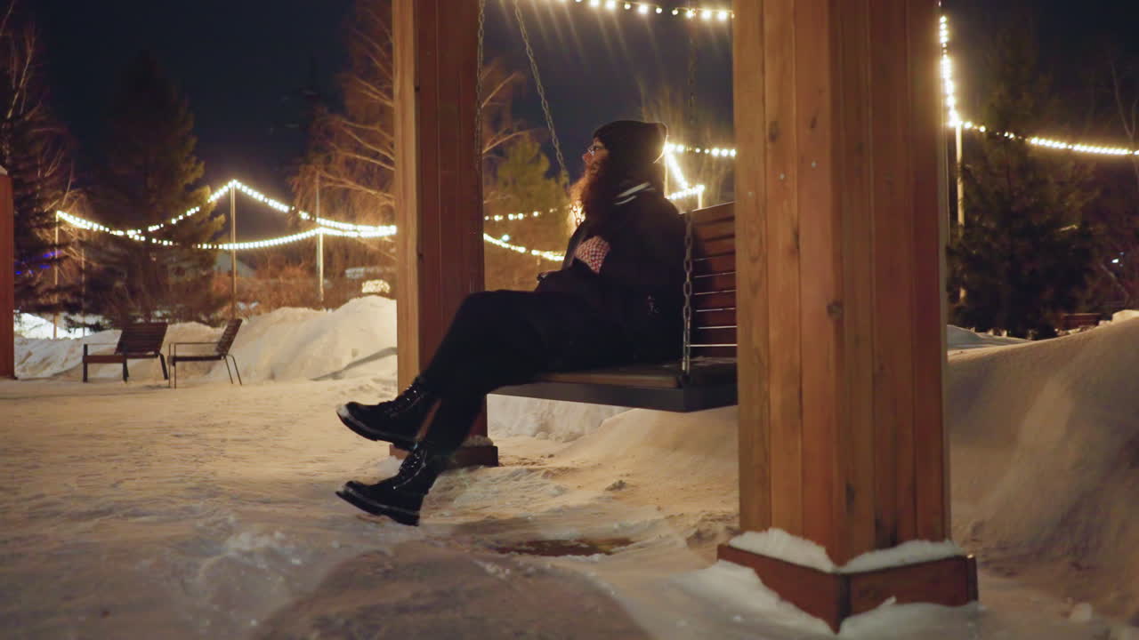 Woman in winter coat and knit hat relaxing on wooden swing under festive string lights at snowy park during cold evening, enjoying peaceful atmosphere surrounded by snowdrifts and warm illumination