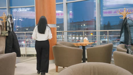 woman in white shirt walks to restaurant table, places coffee and phone on surface, then gracefully sits down on cushioned chair with crossed legs in warm interior with window view of evening city