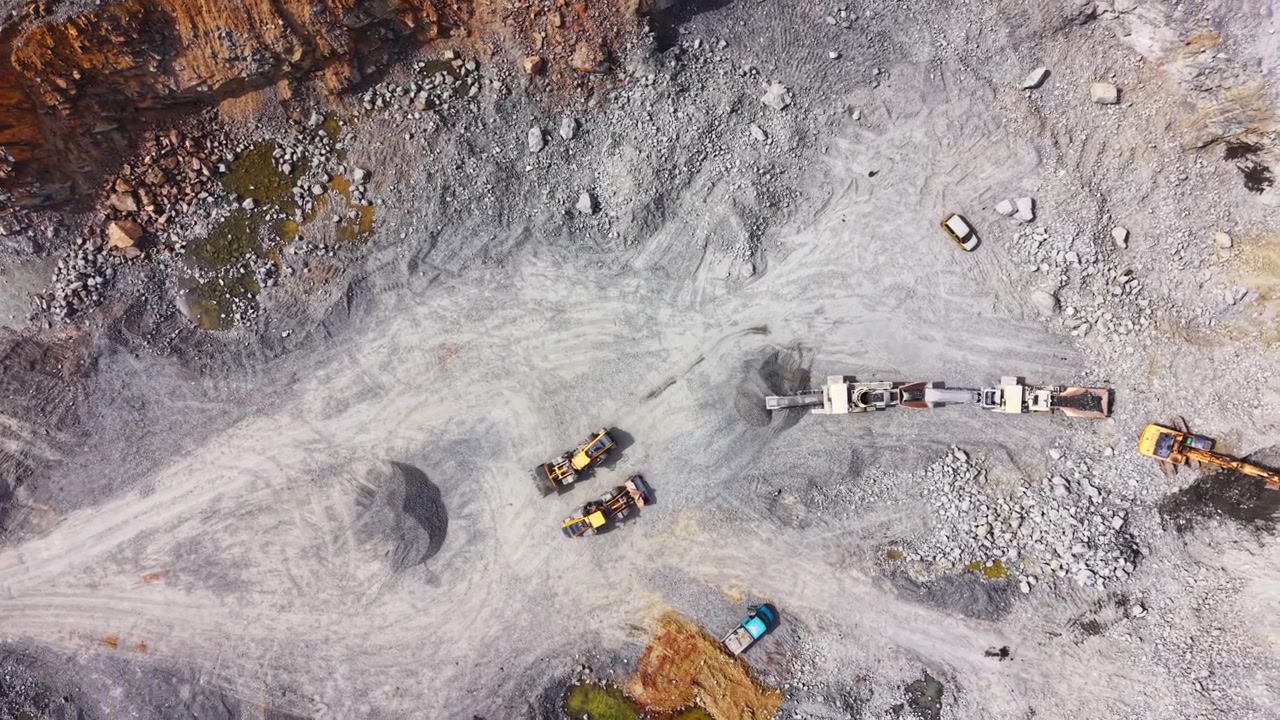 Top down drone view of multiple excavators and mobile crushing units operating on the rocky pit floor. Overhead shot of heavy machinery in a stone quarry.