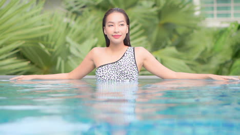 woman relaxing in luxury hotel lounge inside swimming pool leaning on the edge with tropical palm leaves on background - slow-motion