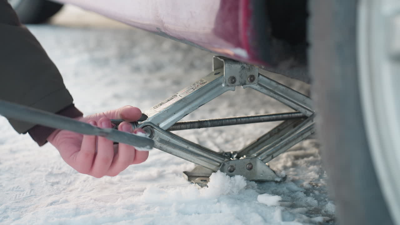 Close up of car mechanic using spanner to turn jack screw and increase height while working under parked vehicle on snowy ground, hands adjusting metal parts